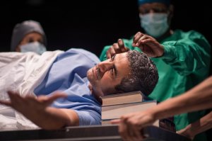 A man in a hospital gown resting his head on a pile of books.  Behind him, a surgeon wearing hospital scrubs and a mask is miming a brain operation. 